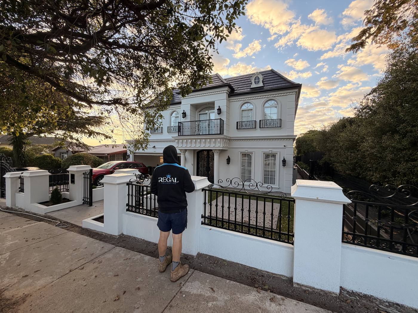 Full-body on the path at a grand white home with iron fence